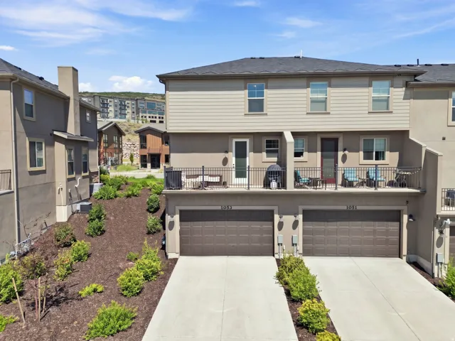 Two-car garage with individual doors, concrete driveways, upper-level balcony with metal railing, and a sloped, landscaped terrain