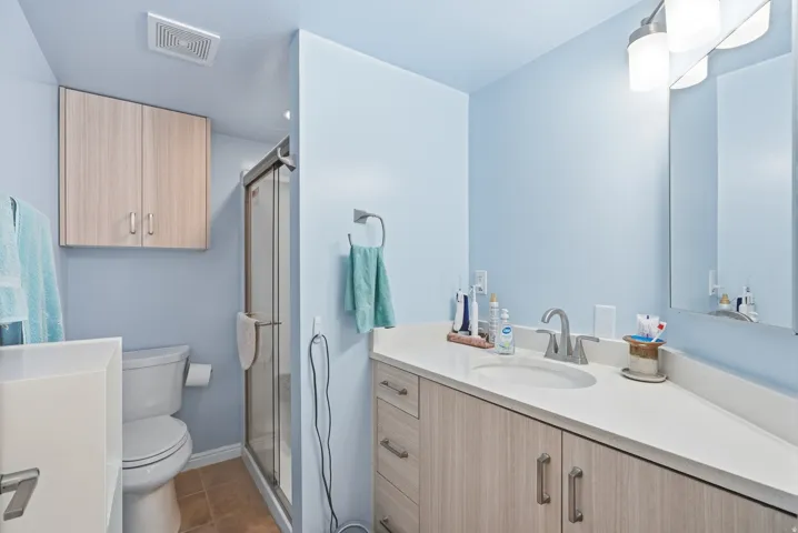 Glass-enclosed shower, light wood-finish cabinetry, white countertop, integrated sink, and wall-mounted vanity lighting