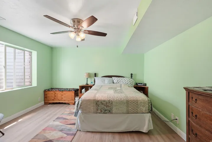 Wood-finish flooring, overhead ceiling fan with light fixture, window with horizontal grates, and light green wall paint