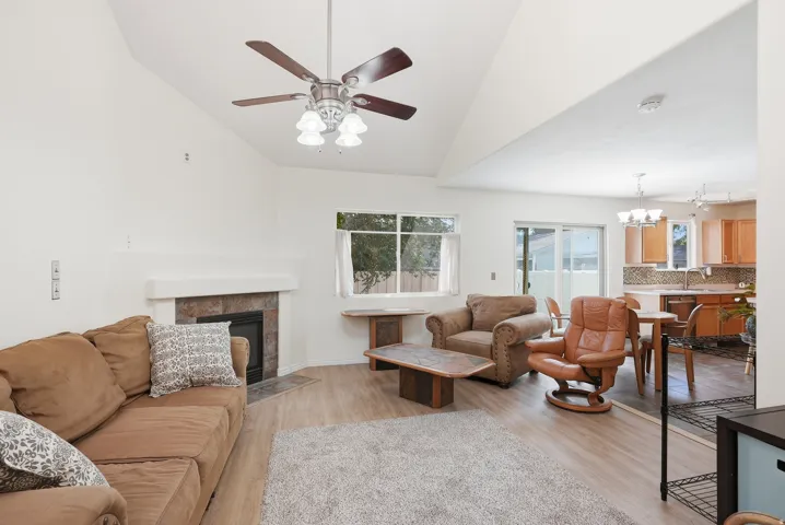 Vaulted ceiling with a ceiling fan, stone-surround fireplace, wood-finish flooring, sliding glass door, and a kitchen with wood cabinetry