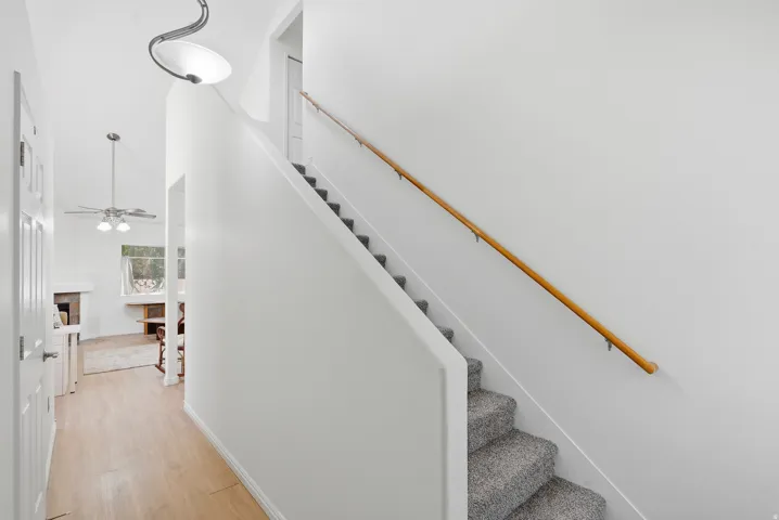 Carpeted staircase with a wood handrail, light wood-finish flooring, a ceiling fan with integrated lighting, and a fireplace with a stone surround