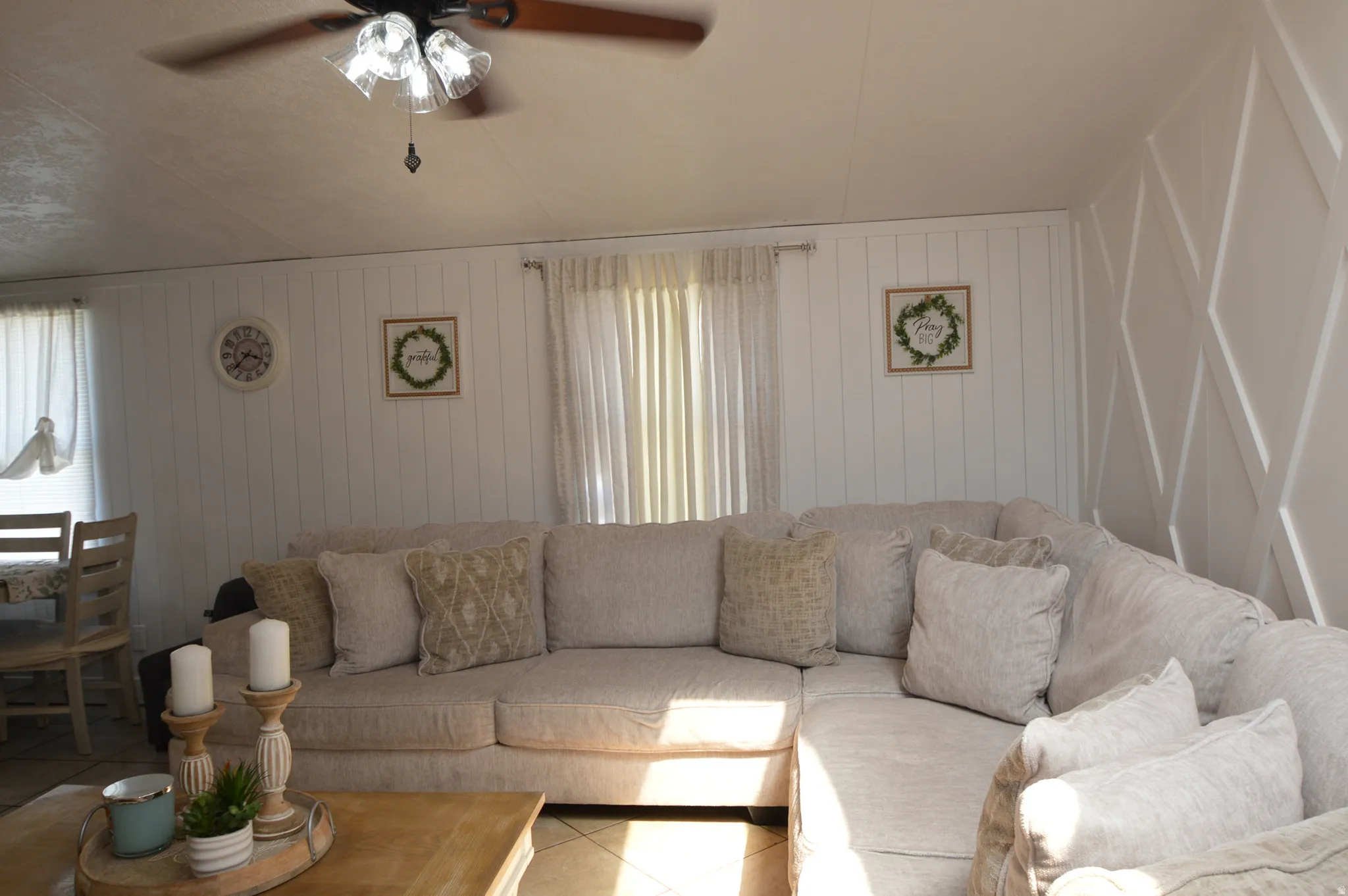 White shiplap accent wall, decorative paneling, ceiling fan with light fixture, tiled flooring, and window with sheers