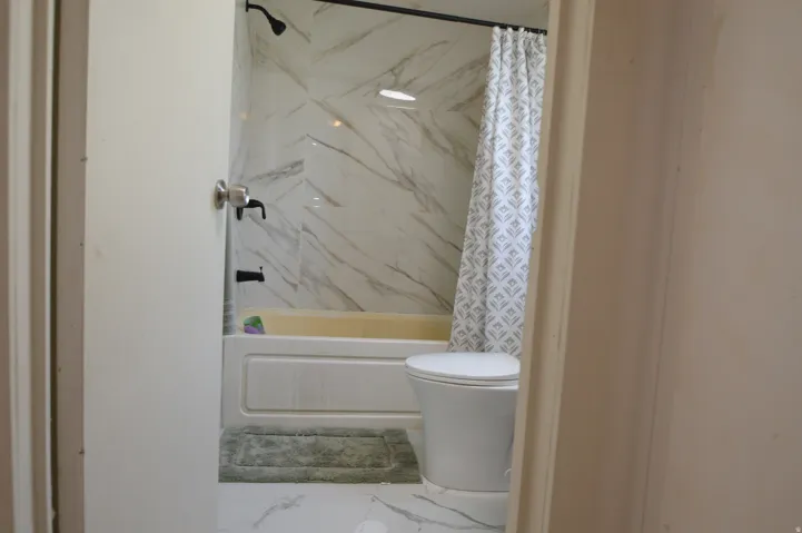 Shower/tub combination with black fixtures, tile surround, white toilet, and tile flooring
