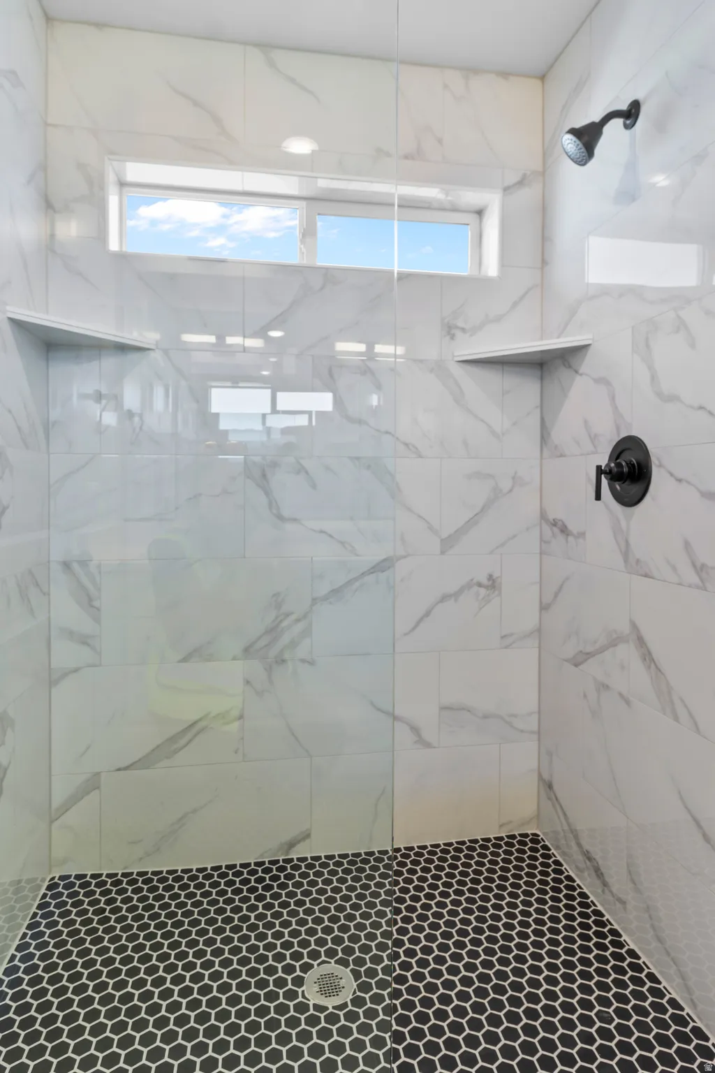 Walk-in shower with full height white tile walls, black hexagonal mosaic tile flooring, built-in shelving, and a transom window