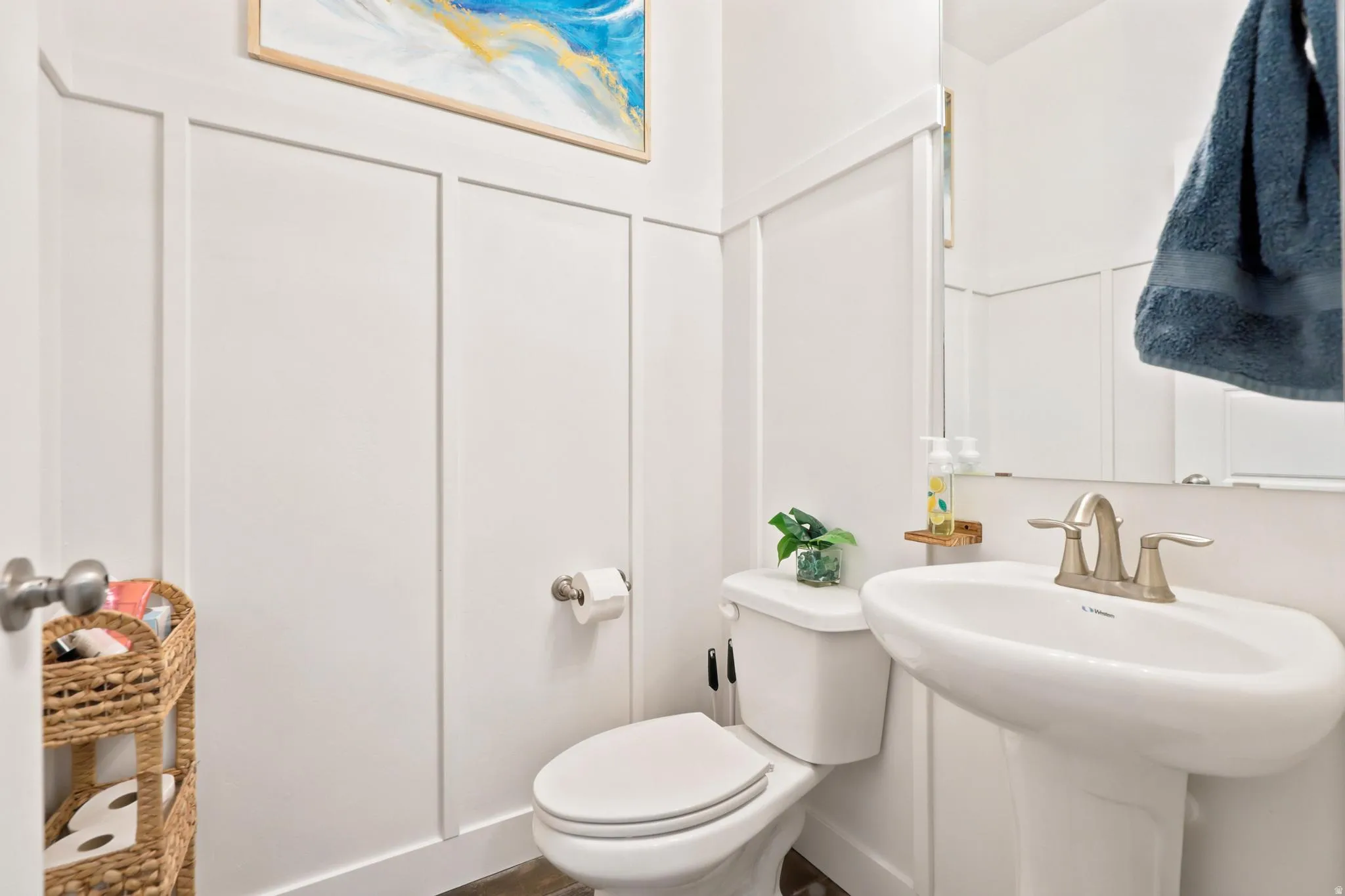 White wainscoting with board and batten detailing, pedestal sink with a two-handle faucet, wall-mounted mirror, and wood-finish flooring