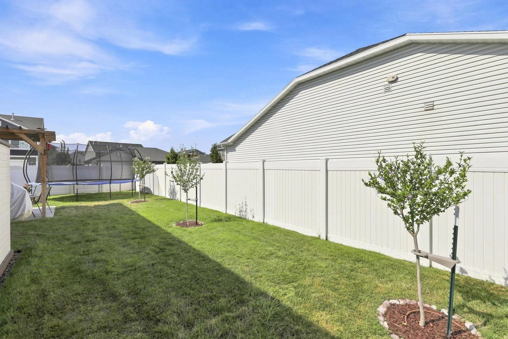 Expansive backyard featuring a manicured lawn, established trees, a white vinyl privacy fence, and a concrete patio