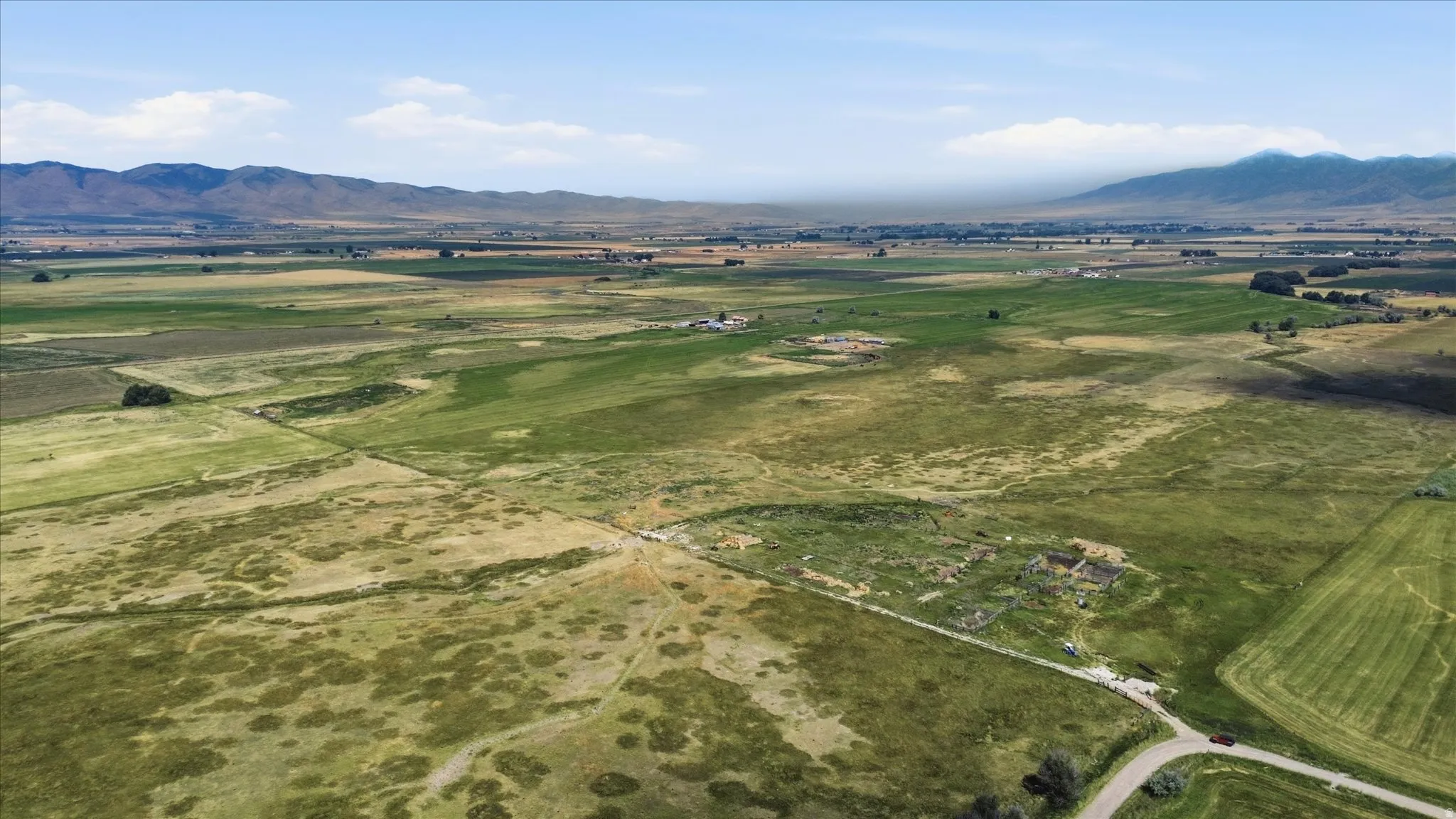 Expansive land featuring green pastures, a winding road, and distant mountain ranges under a clear sky