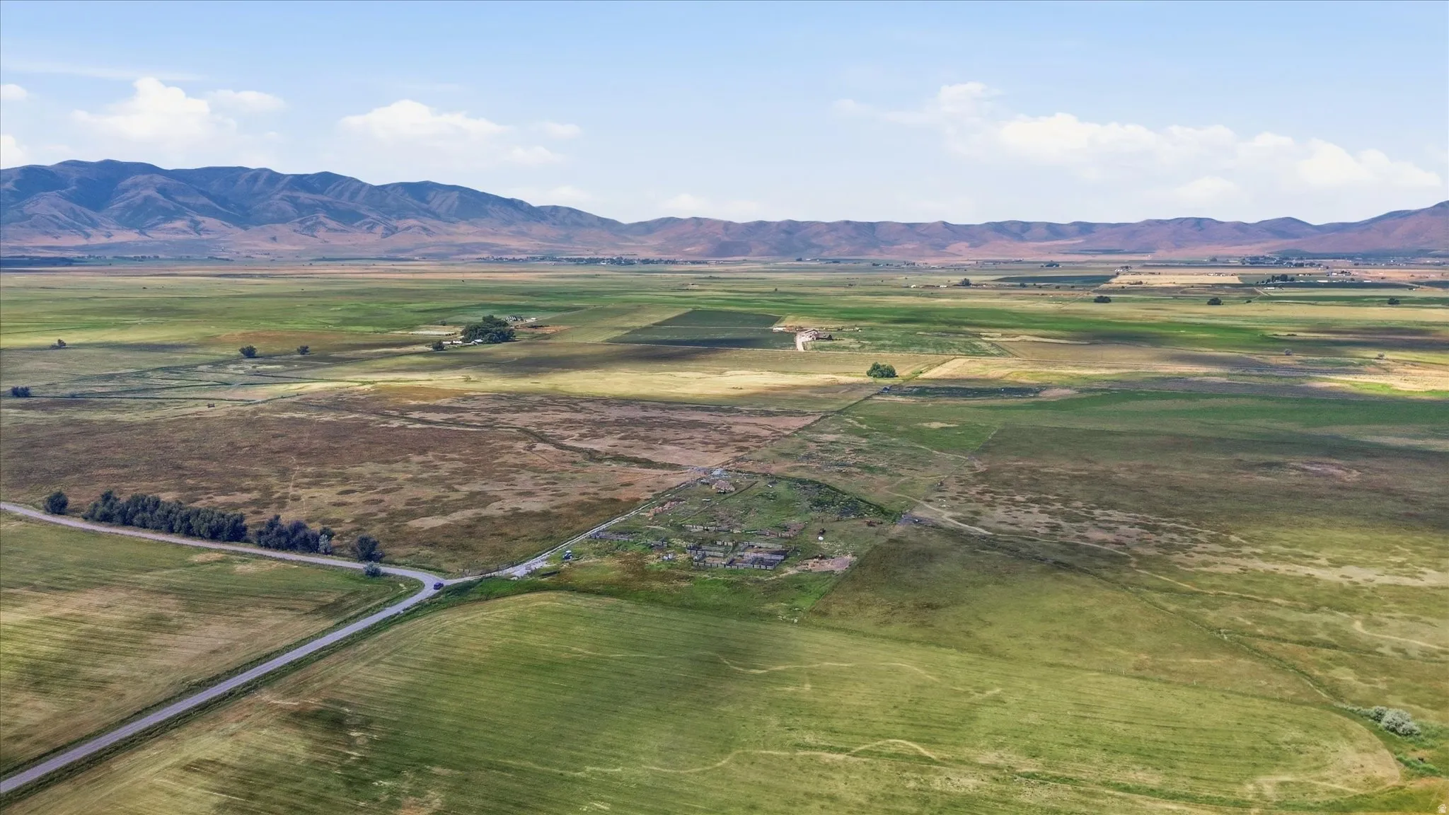 Expansive agricultural land featuring a paved road, scattered trees, distant structures, and a prominent mountain range backdrop