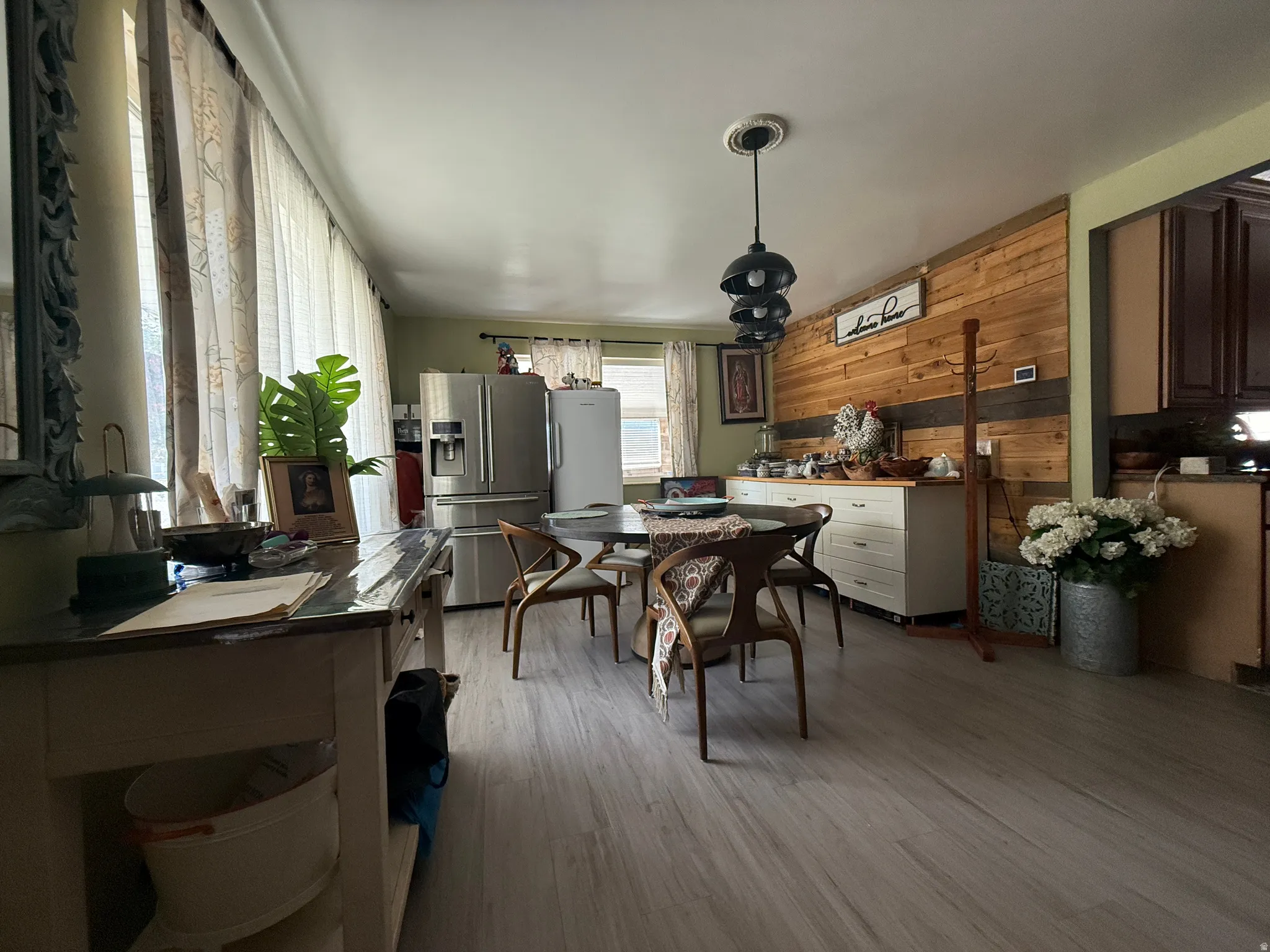 Light wood-finish flooring, two large windows with curtain rods, black multi-pendant light fixture, wood plank accent wall, and built-in white cabinetry with wood countertop