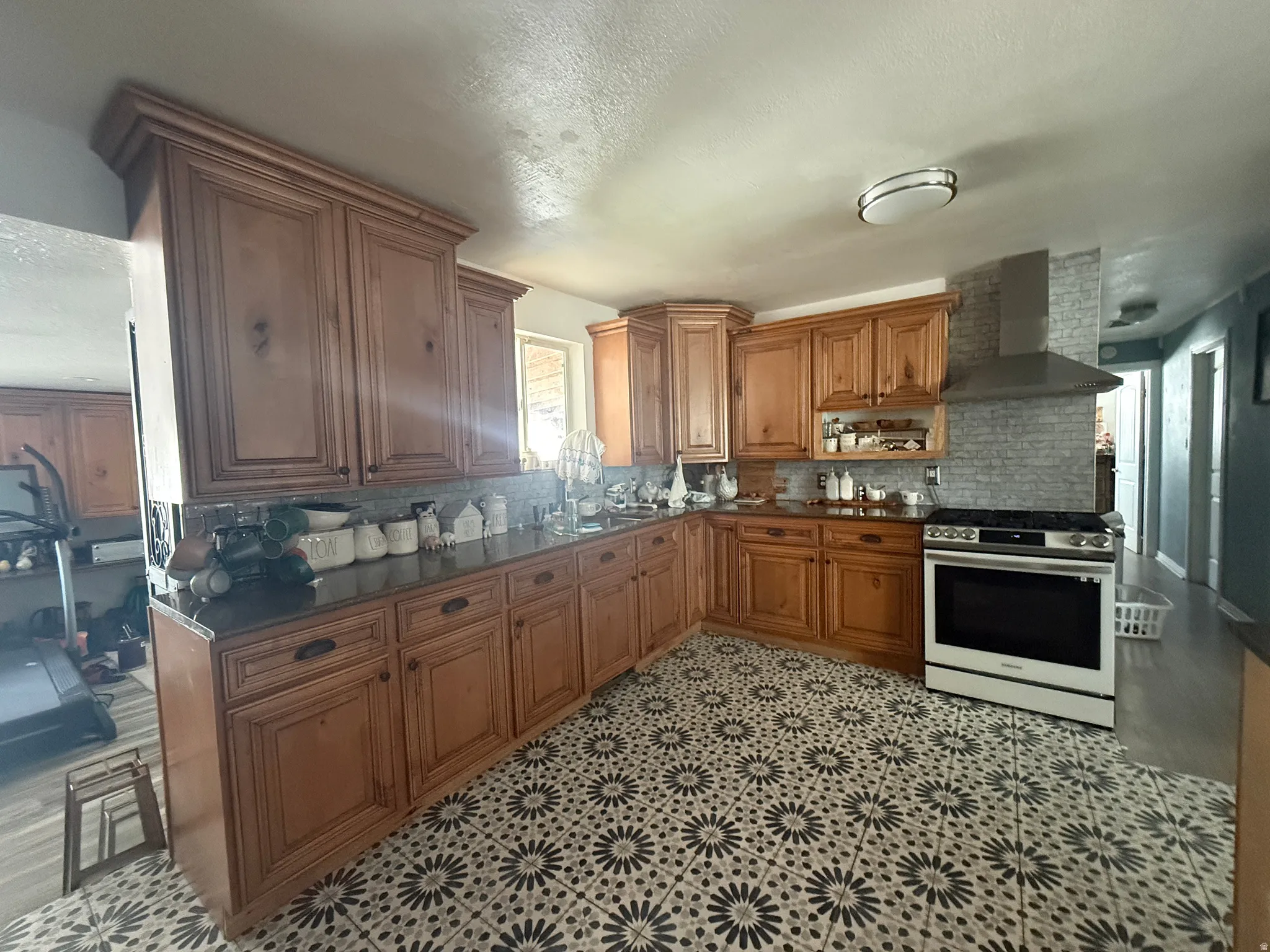 Wood-finish cabinetry, dark countertops, patterned floor tile, stainless steel range with hood, and a tiled backsplash