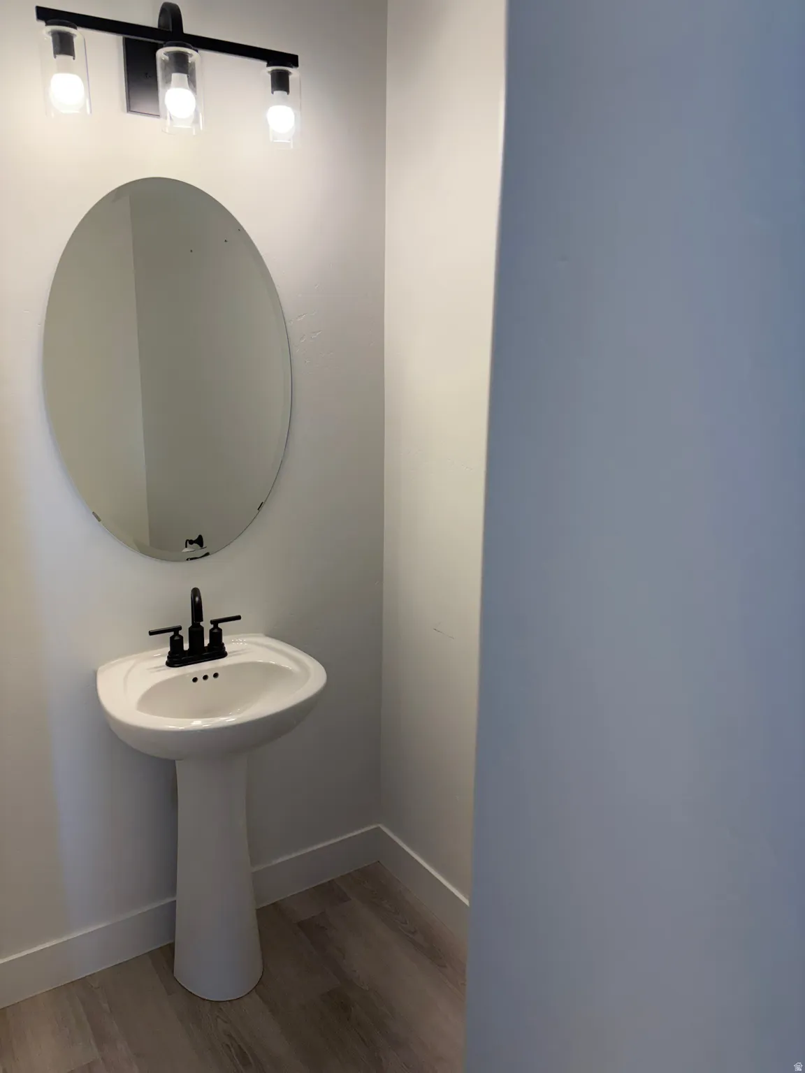 Pedestal sink with a matte black faucet, wall-mounted oval mirror, and wood-finish flooring
