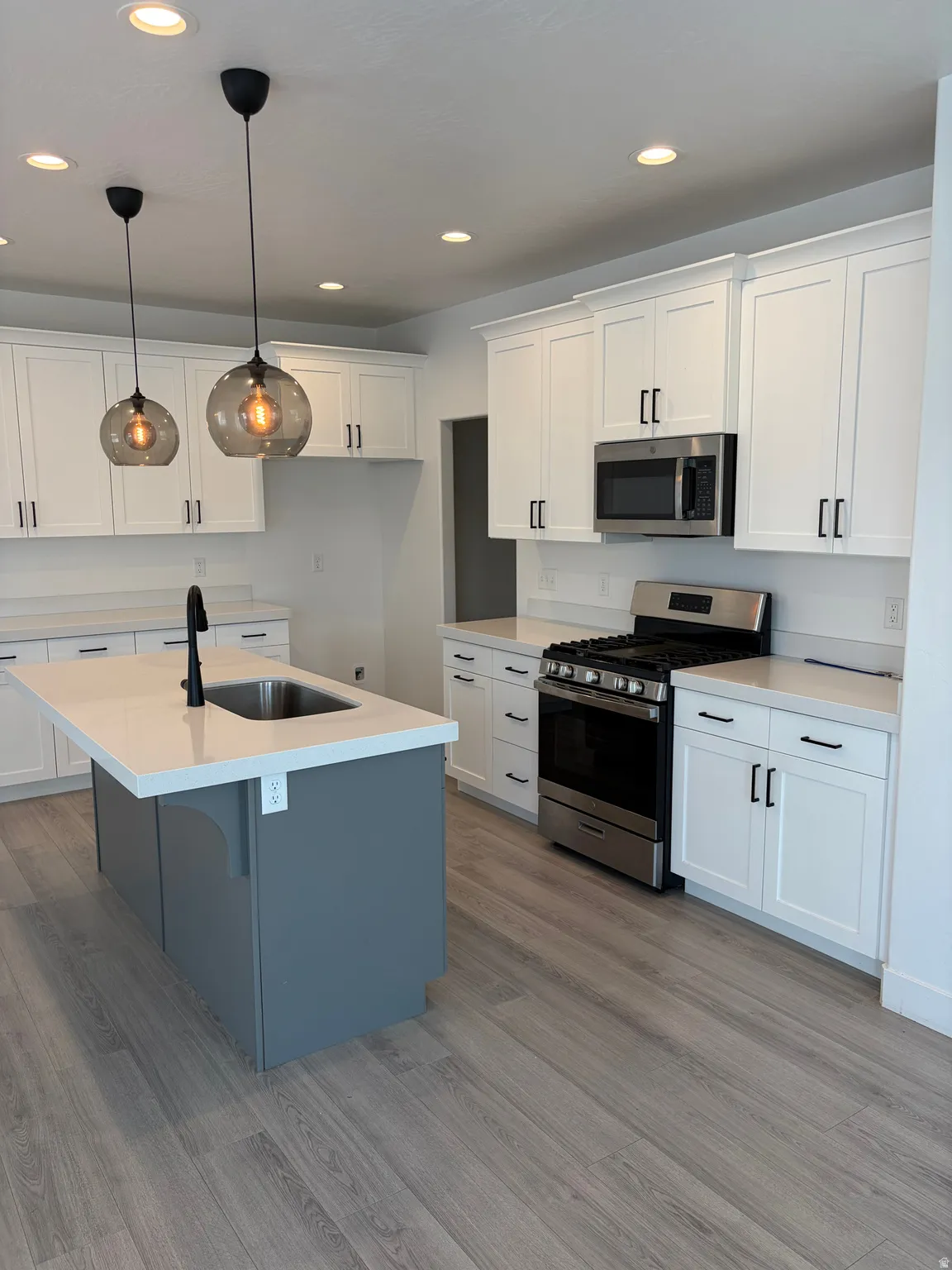 White shaker cabinetry, light-toned wood-finish flooring, stainless steel appliances, light-colored countertops, and a kitchen island with a built-in sink