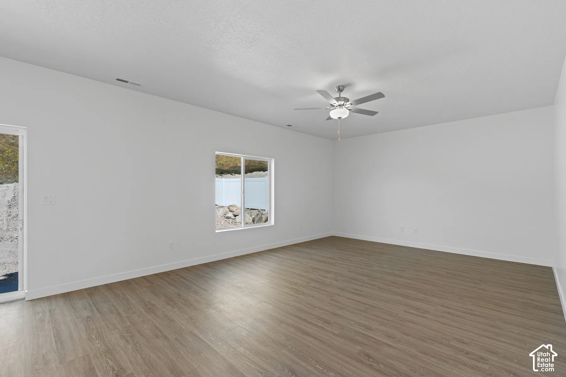 Spacious room featuring wood-finish flooring, white walls, a ceiling fan with integrated lighting, and a large window