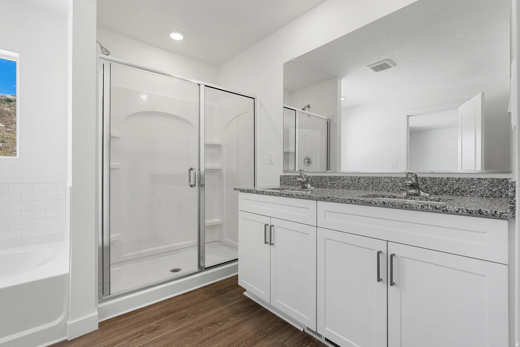 Dual vanity with granite-style countertop, white cabinetry, wood-finish flooring, glass-enclosed shower, and built-in soaking tub