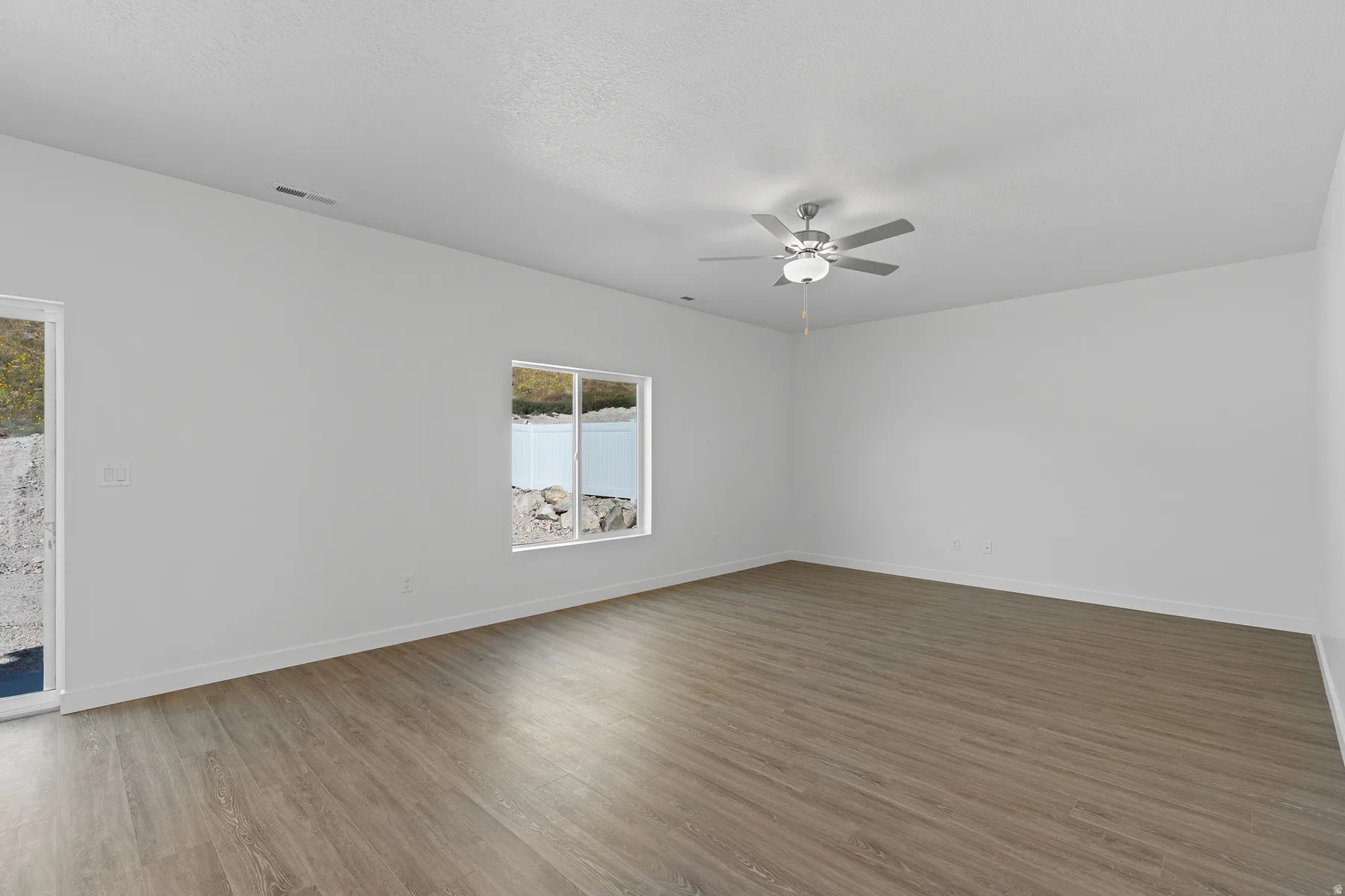 Spacious room featuring wood-finish flooring, white walls, a ceiling fan with integrated lighting, and a large window