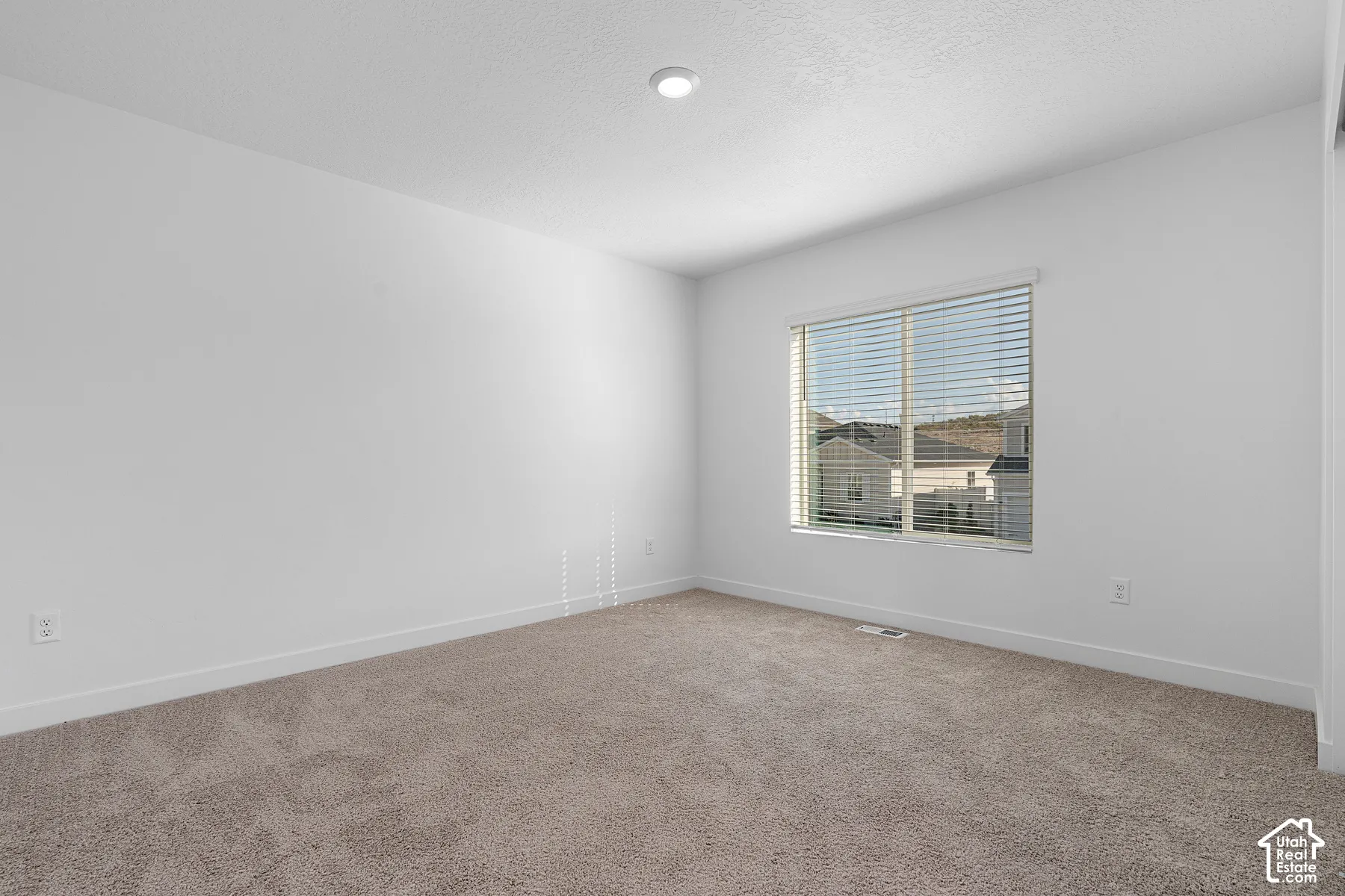 Bright bedroom featuring neutral carpet flooring, recessed ceiling lighting, and large double-hung window with horizontal blinds offering natural illumination