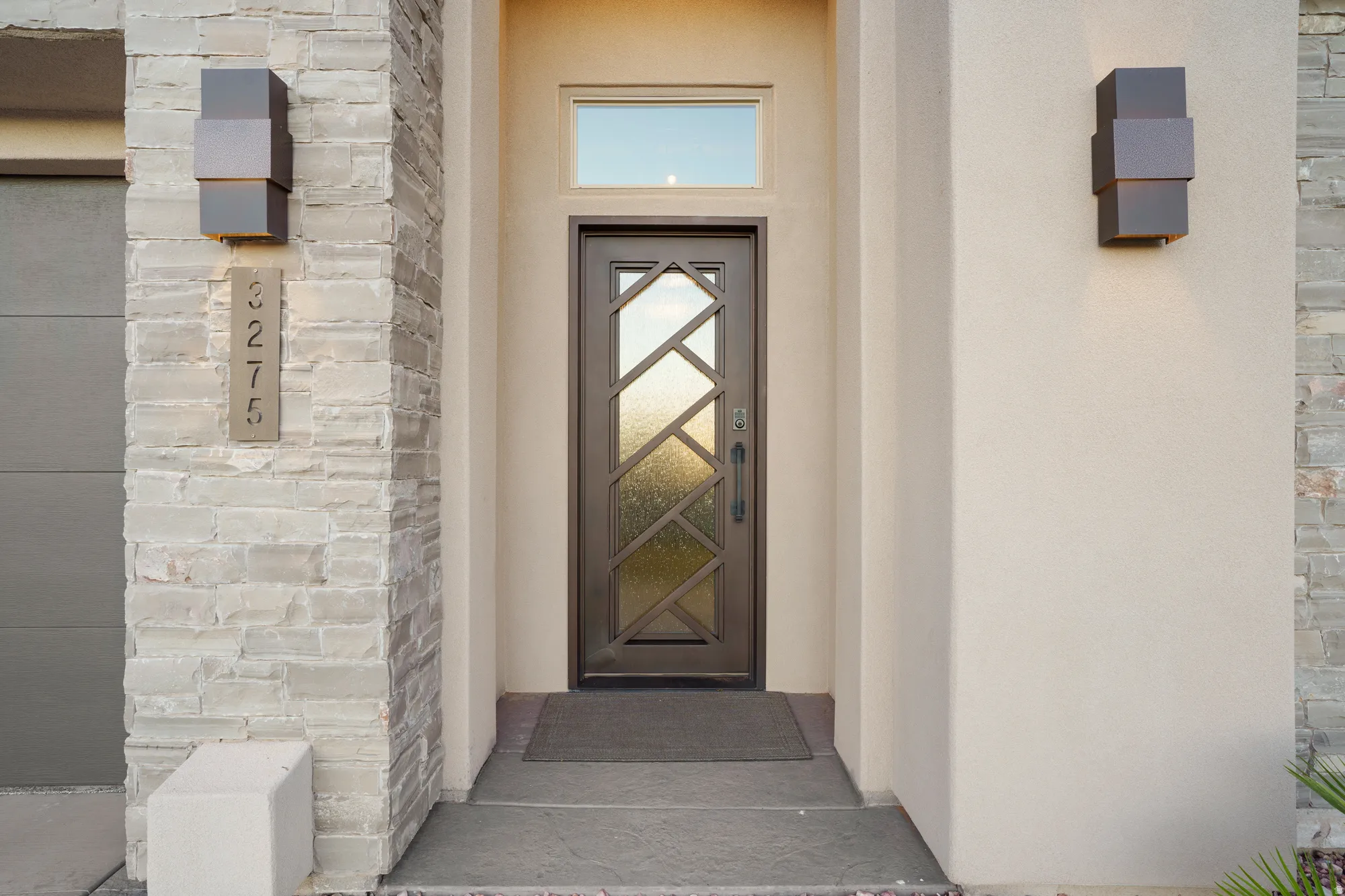 Entryway featuring a decorative transom window, a bronze door with geometric glass inserts, stucco siding, stacked stone accents, and modern exterior lighting
