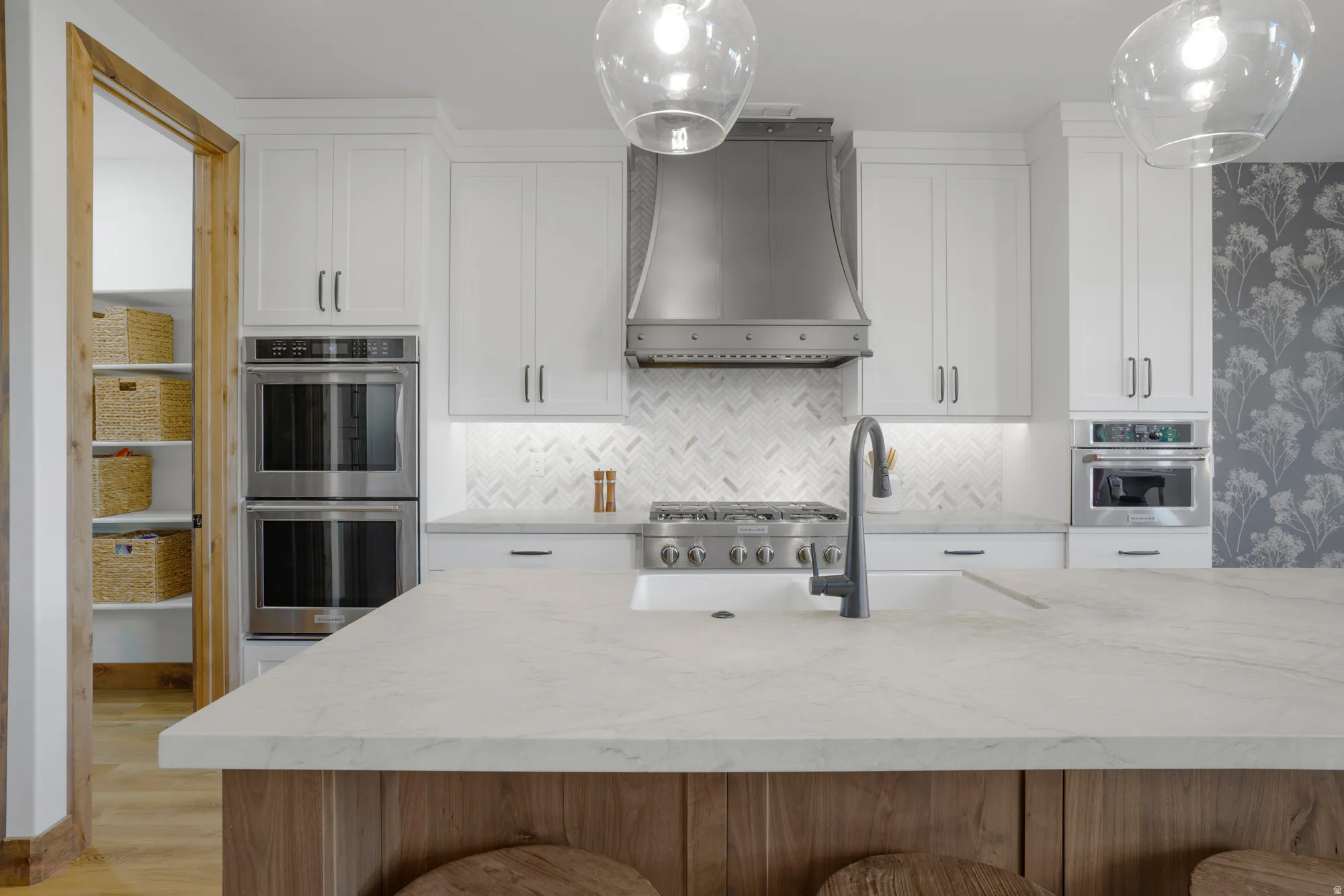 White shaker cabinetry, stainless steel double ovens, a herringbone tile backsplash, a gas range, and a large island with a surface-mounted sink
