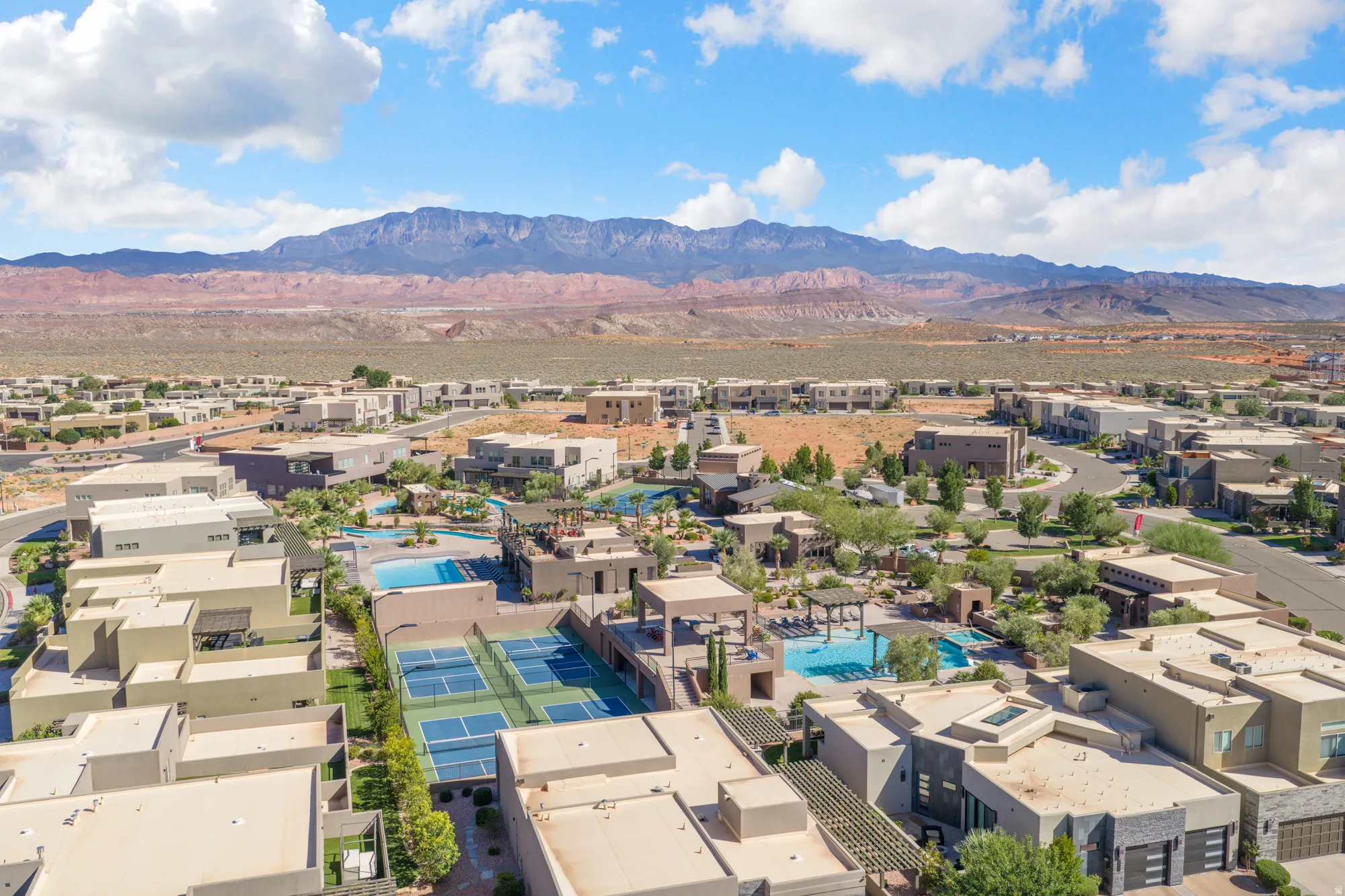 Expansive community featuring multiple tennis courts, swimming pools, and residential structures with flat roofs, all set against a mountainous backdrop