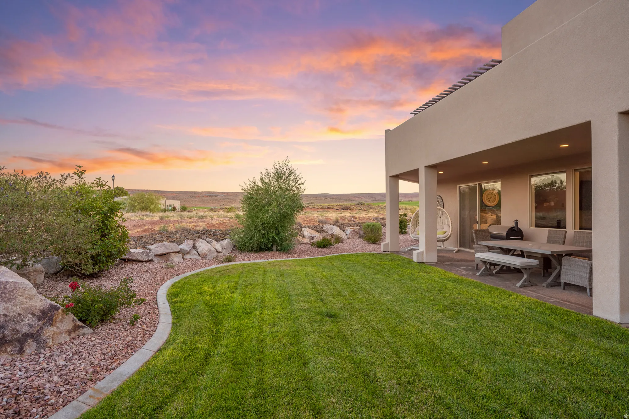 Expansive lawn bordered by a rock-lined garden bed, covered patio with recessed lighting, and sliding glass door access