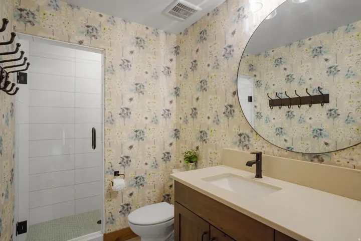 Frameless glass shower enclosure with white tiled walls, vanity with integrated sink, dark bronze-finish fixtures, and a large circular mirror