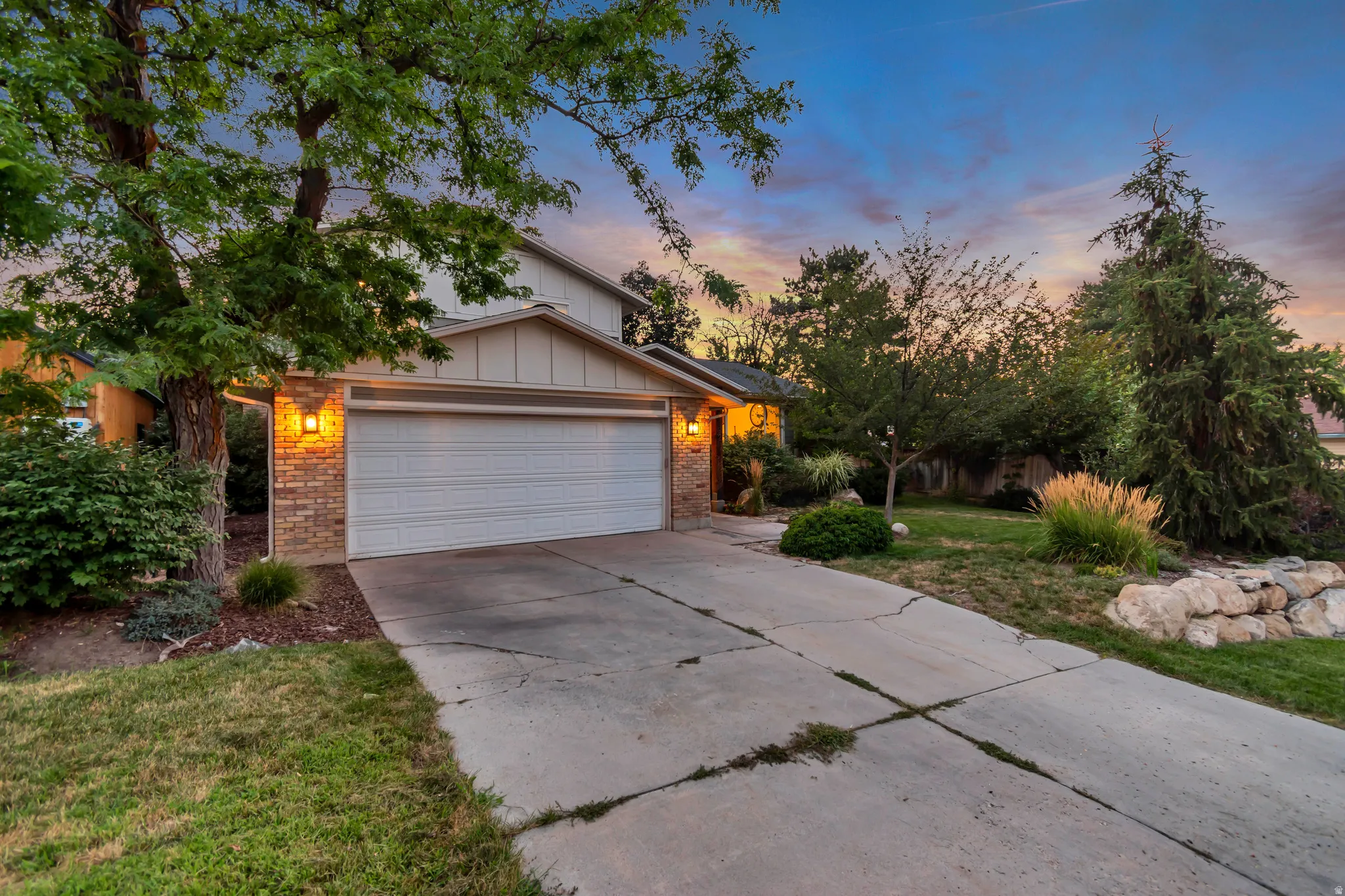 Attached two-car garage, brick facade detailing, mature landscaping, exterior wall sconces, and a concrete driveway
