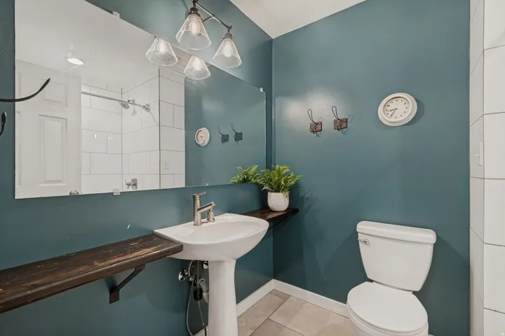 Pedestal sink with a single-handle faucet, vanity mirror, wall-mounted light fixture, white toilet, and tile flooring