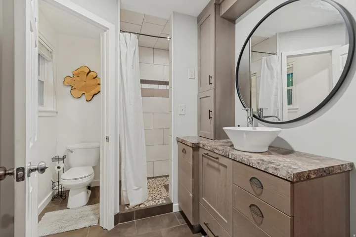 Vanity with vessel sink and round mirror, complementing a tiled shower with a stone floor, and a toilet alcove with a window