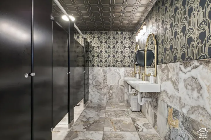 Spa-inspired bathroom featuring a spacious walk-in shower with black framed glass enclosure, dual vanity with polished stone countertops, decorative tile wainscoting, and ornate patterned wallcovering