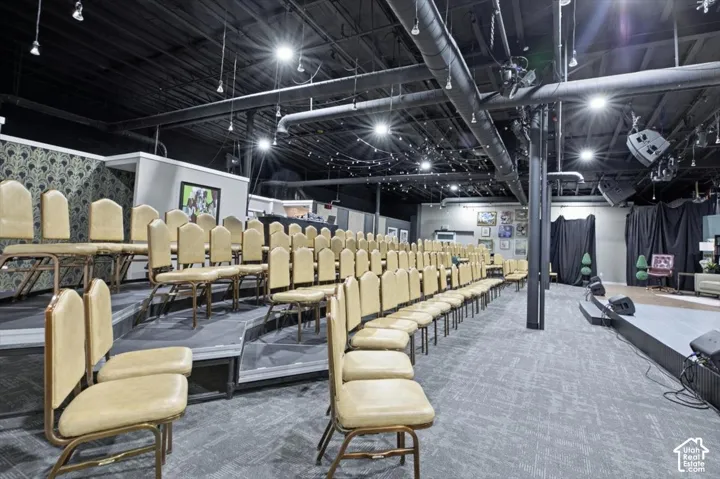 Exposed industrial-style ceiling with track lighting, elevated tiered seating, carpeted flooring, and a raised stage area
