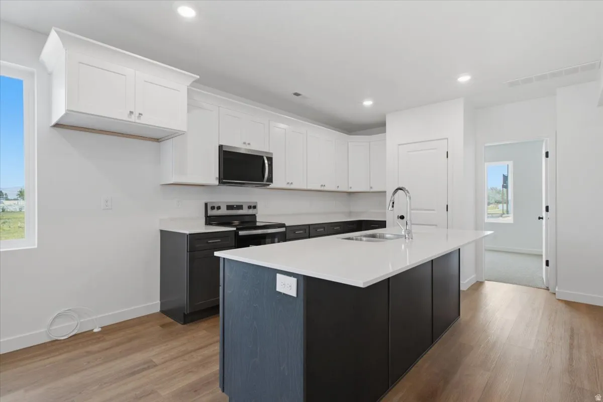 Kitchen featuring white upper cabinetry, dark lower cabinetry, stainless steel appliances, a center island with sink, and wood-finish flooring
