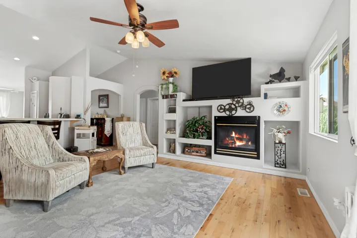 Living area featuring wood-finish flooring, a vaulted ceiling, a ceiling fan with integrated lighting, and a built-in fireplace with shelving