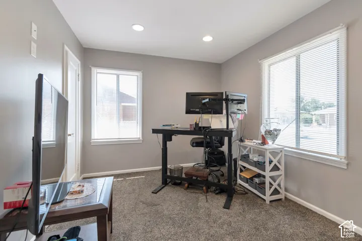Recessed lighting illuminates the space, featuring two windows with white blinds, neutral wall tones, and carpet flooring