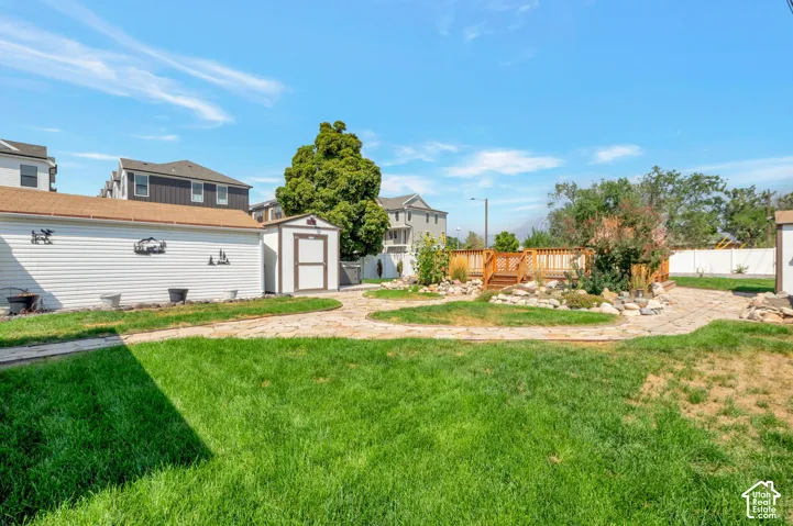 Expansive backyard featuring a manicured lawn, a raised wood deck, paved pathways, and 2 storage sheds with lights and electricity