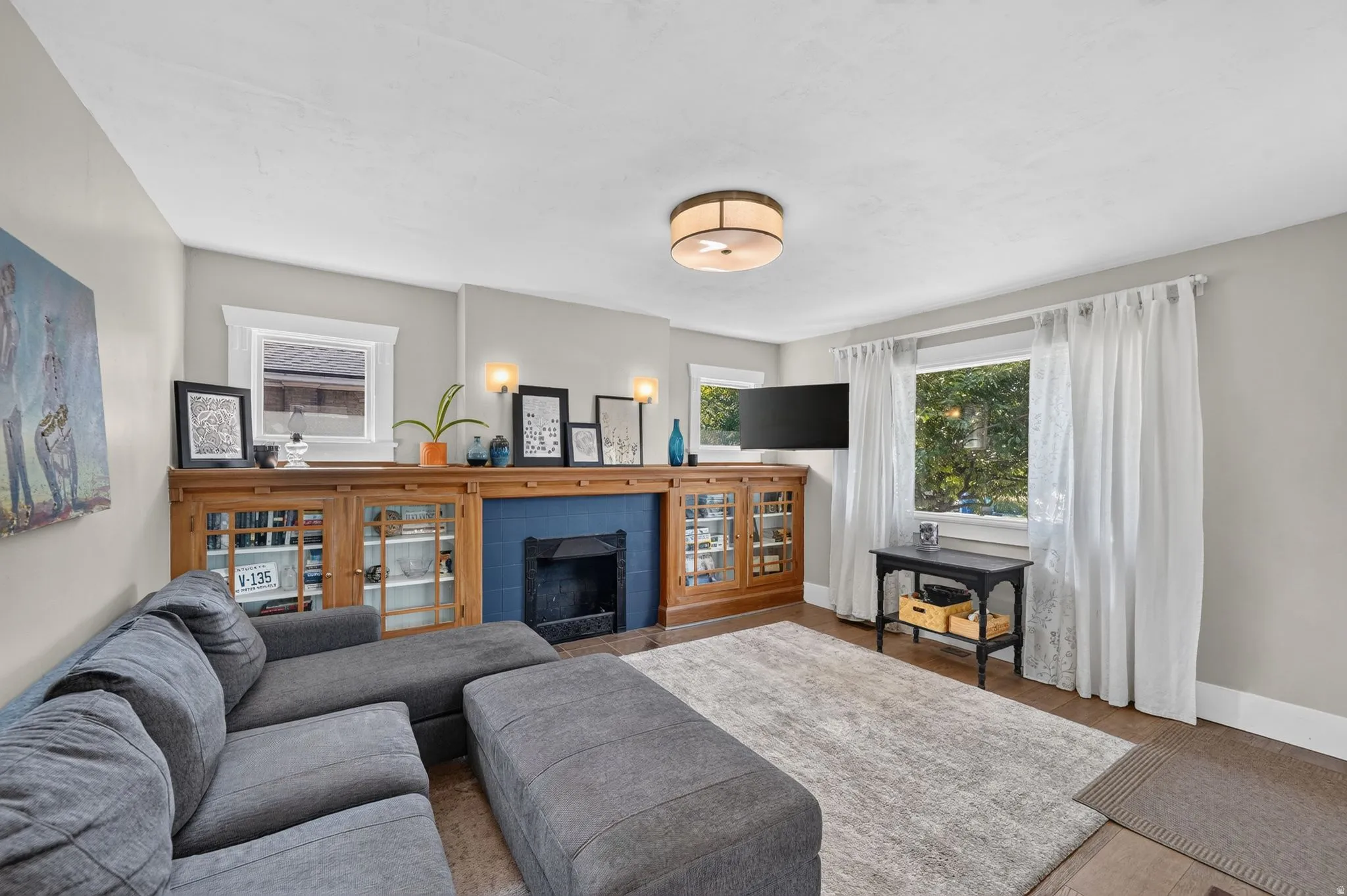 Hardwood flooring, ceiling-mounted light fixture, built-in cabinetry with glass doors, fireplace with blue tile surround, and multiple windows