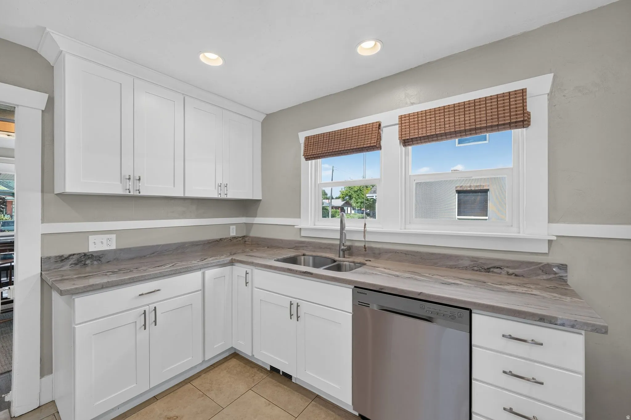 White shaker-style cabinetry, stainless steel dishwasher, double basin sink with gooseneck faucet, light-toned tiled flooring, and recessed lighting