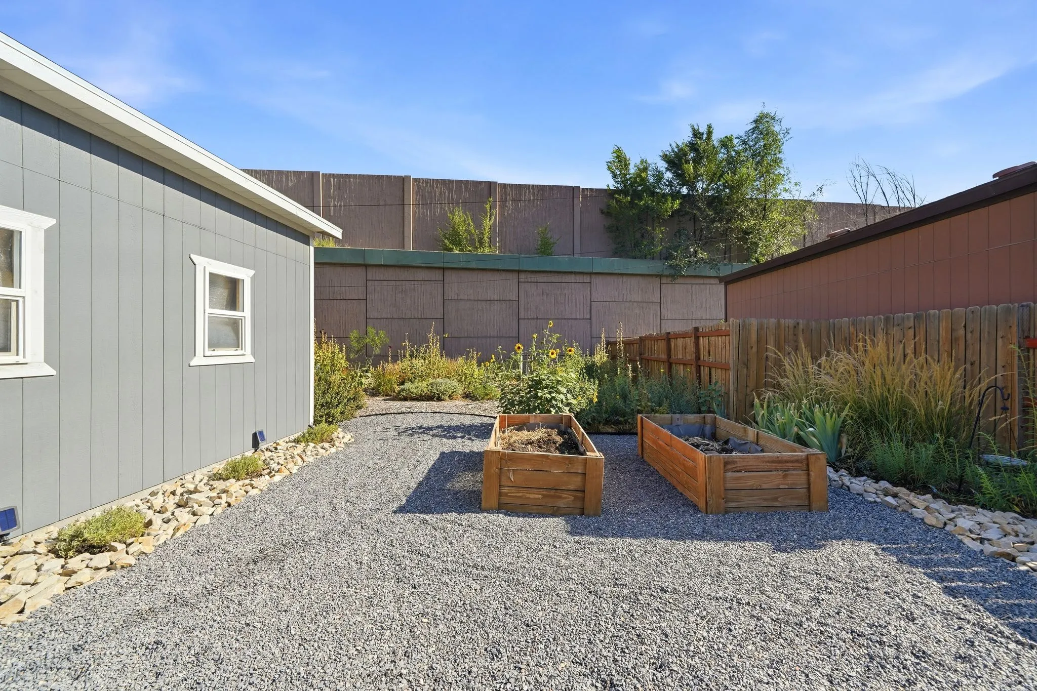 Gravel patio space with two wooden raised garden beds, surrounded by established plantings and a wood fence