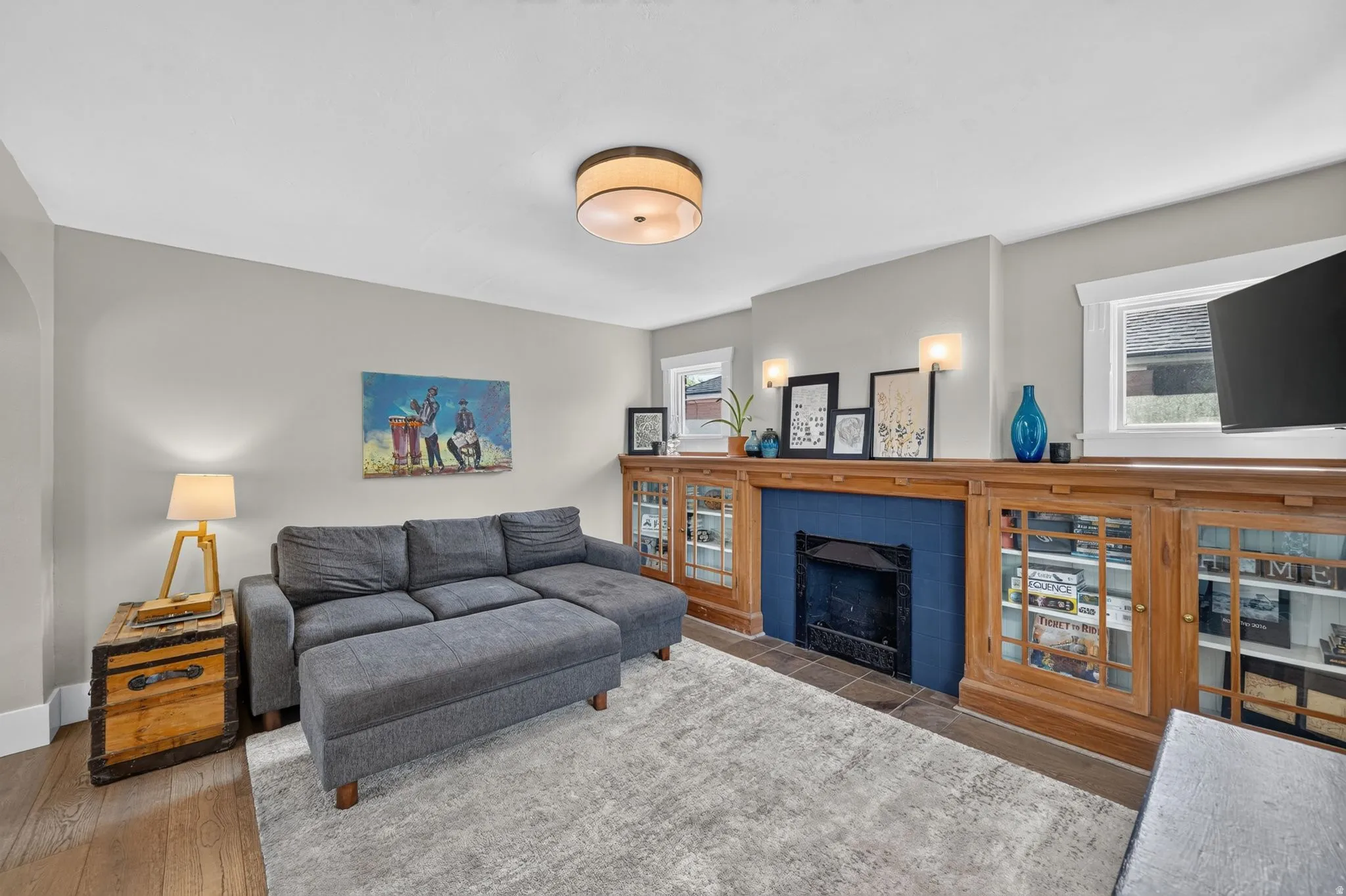 Wood-finish flooring, built-in fireplace with blue tile surround, built-in display cabinetry with glass panel doors, flush-mount ceiling light, and wall sconces