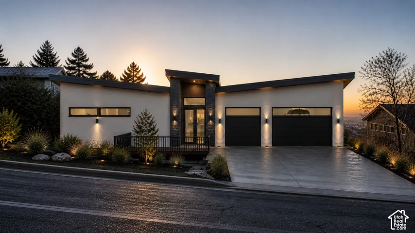 Modern exterior with a sloped roofline, stone accent siding, integrated exterior lighting, a three-car garage, and landscaped grounds