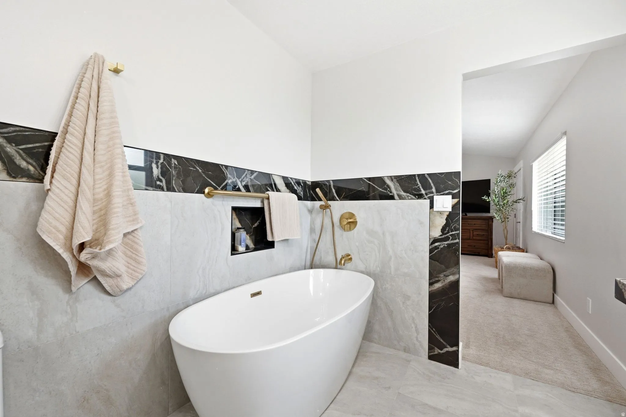 Freestanding soaking tub with gold-tone fixtures, black and gray marble-look tile accents, recessed shower niche, and light-toned floor tile
