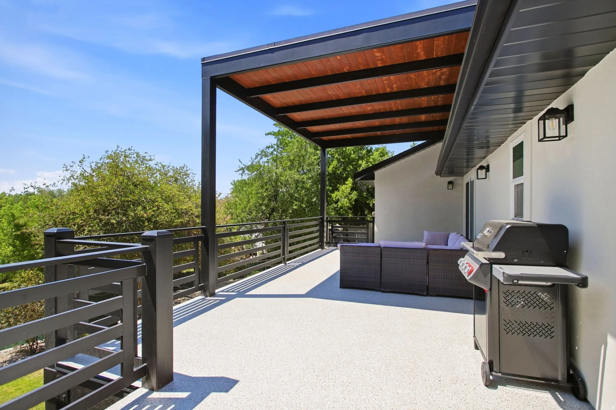 Expansive outdoor deck featuring a covered pergola with a wood-finish ceiling, black metal railings, and exterior wall lighting