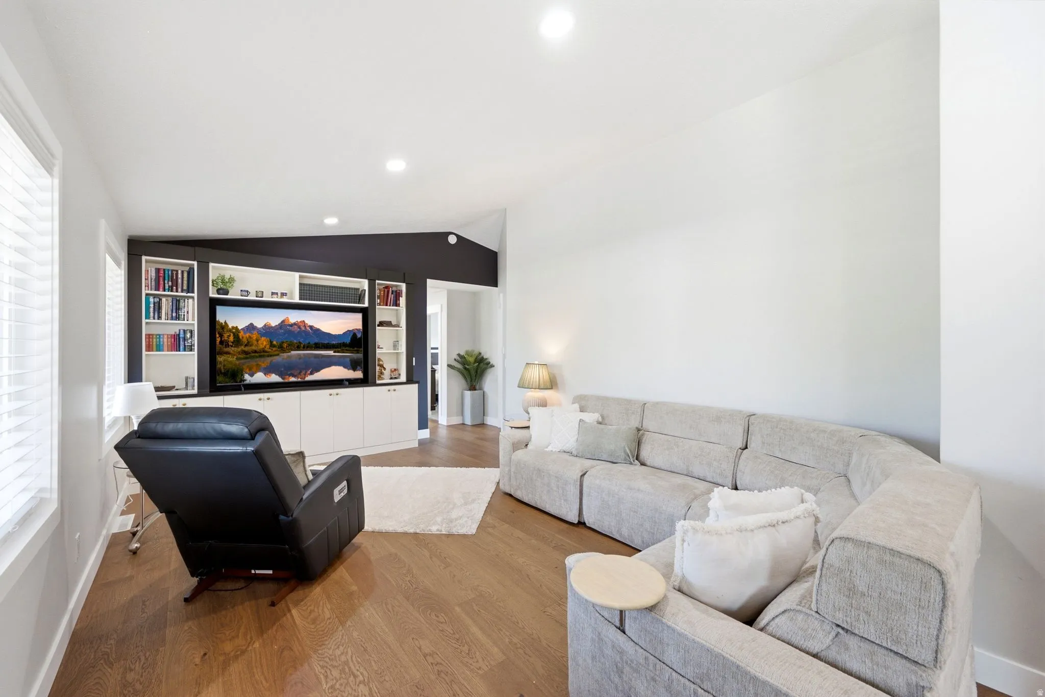 Vaulted ceiling with recessed lighting, wood-finish flooring, built-in media center with shelving, and large window with blinds