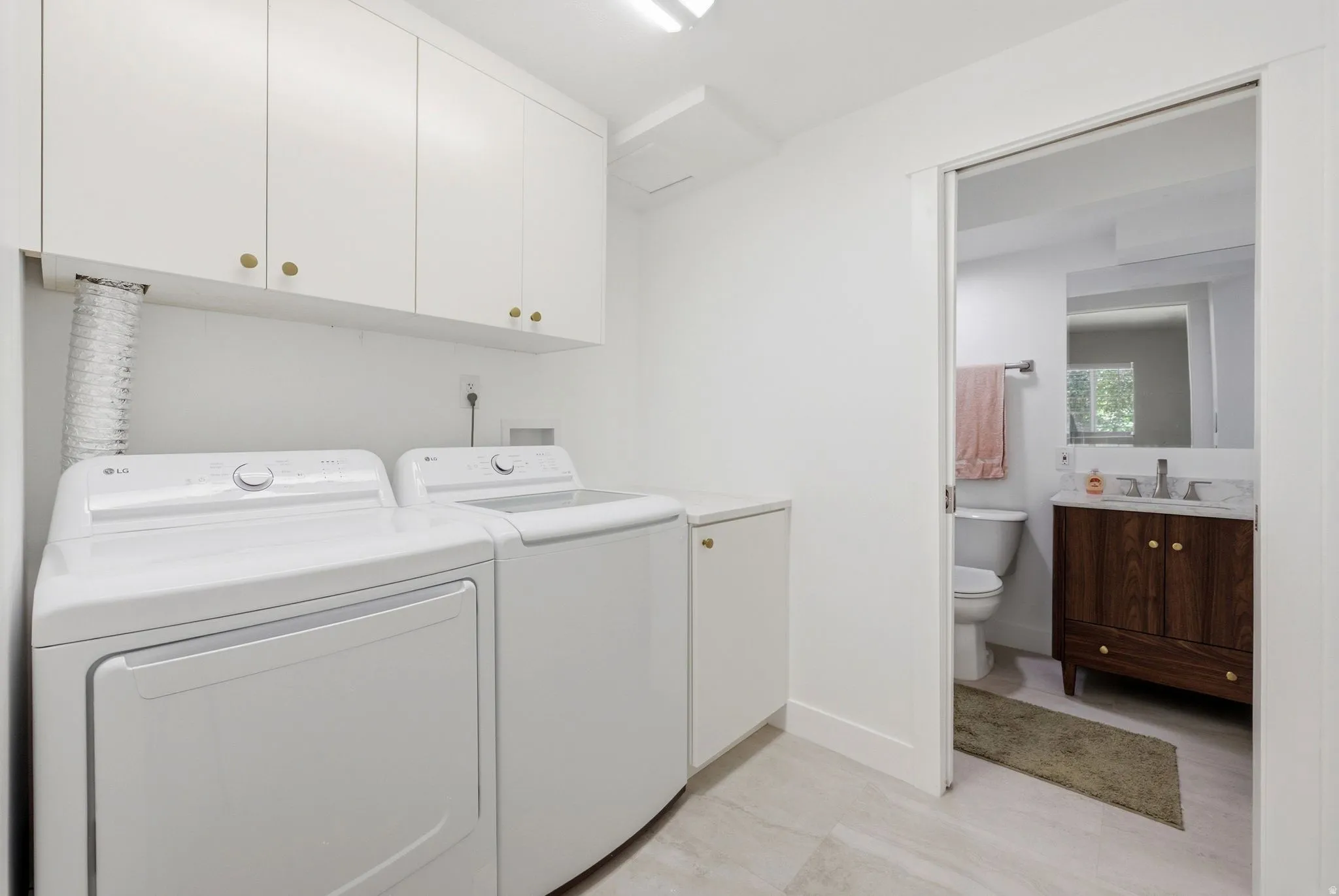 White upper cabinetry with gold-finish hardware, light-toned floor tile, and a sliding pocket door opening to an additional space featuring a wood-finish vanity with a stone countertop