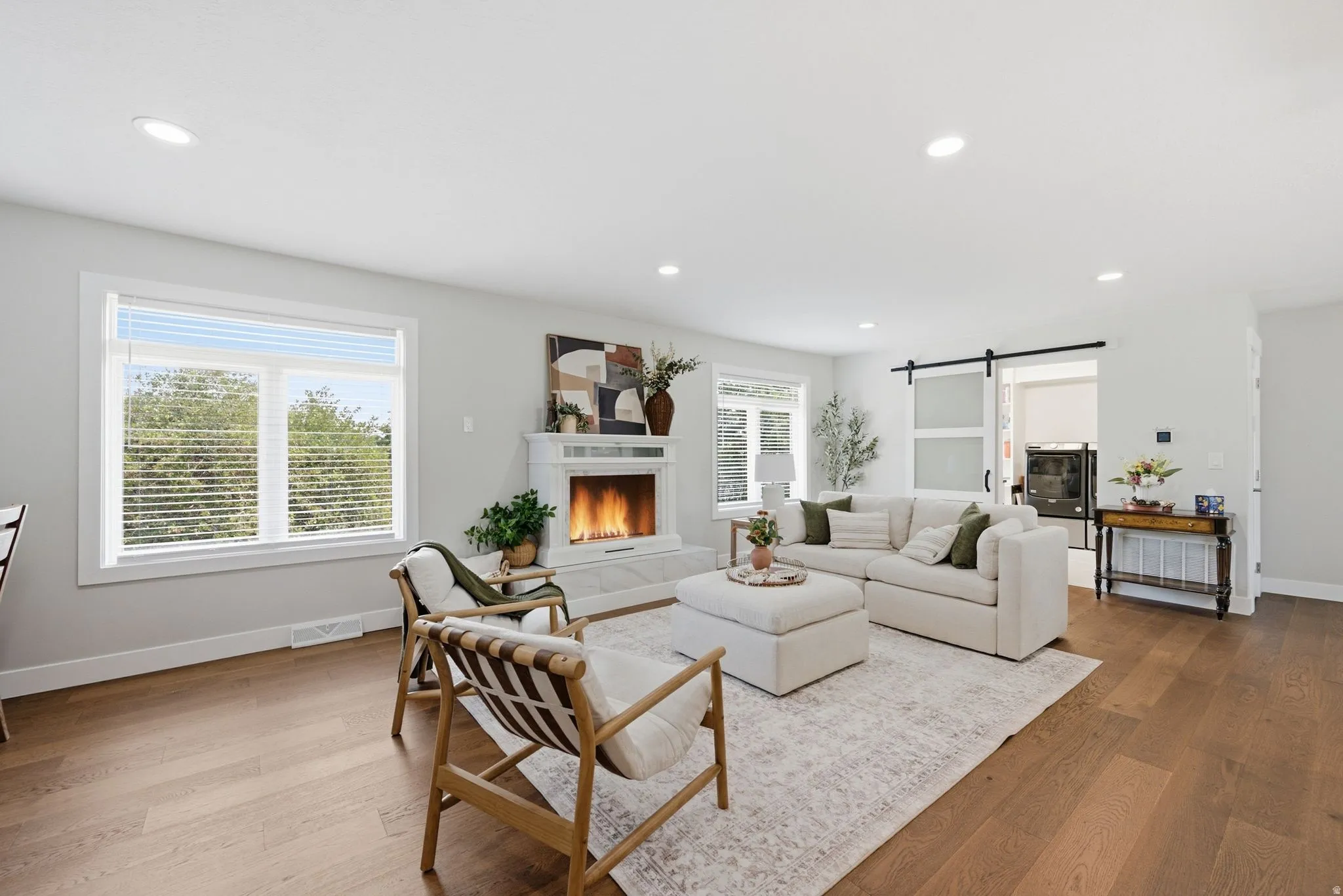 Spacious living area featuring wood-finish flooring, a white mantel fireplace, abundant natural light from large windows, recessed lighting, and a modern barn door