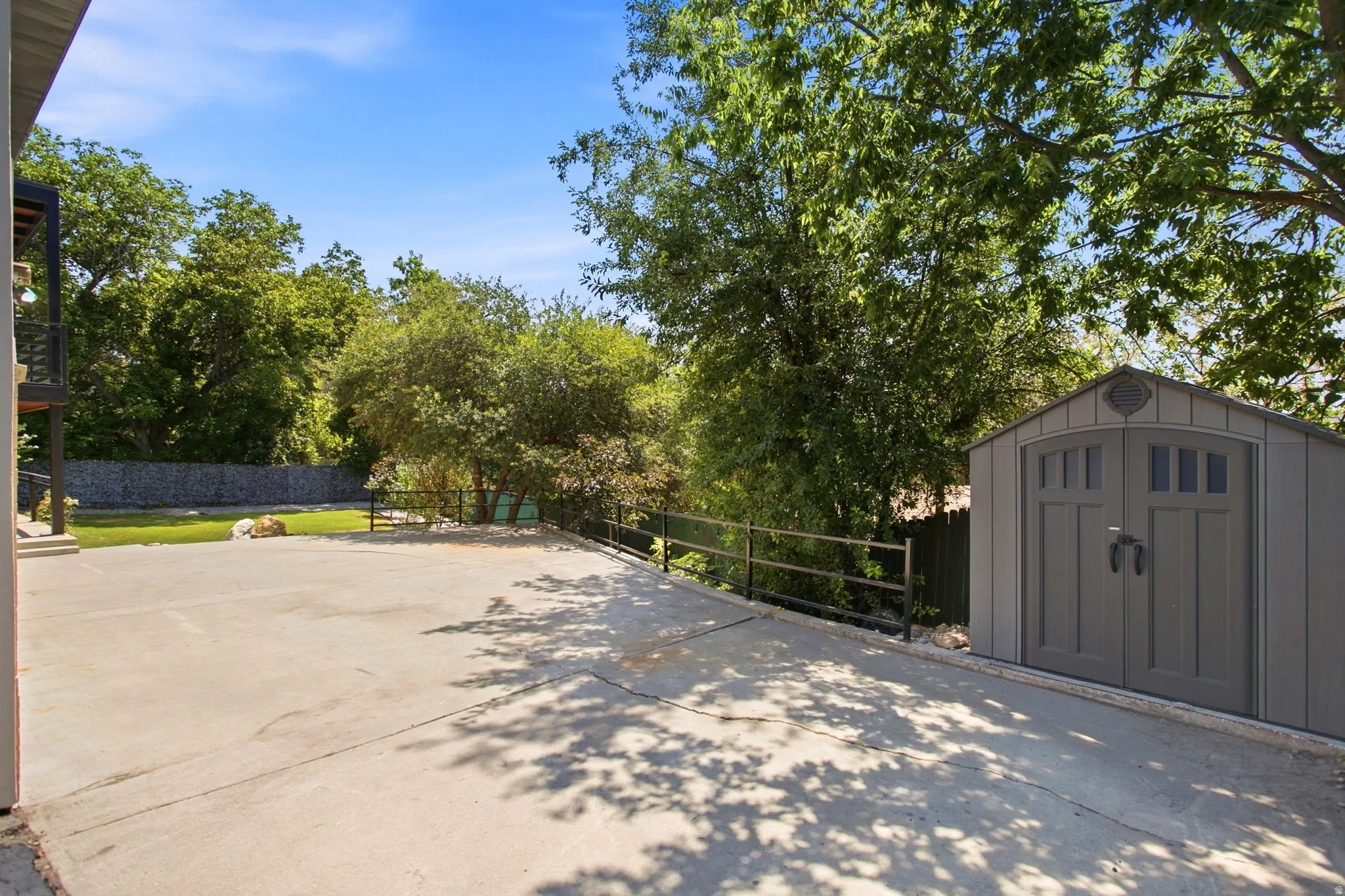 Paved concrete patio area with a detached storage shed featuring double doors, a metal fence, and a stone retaining wall