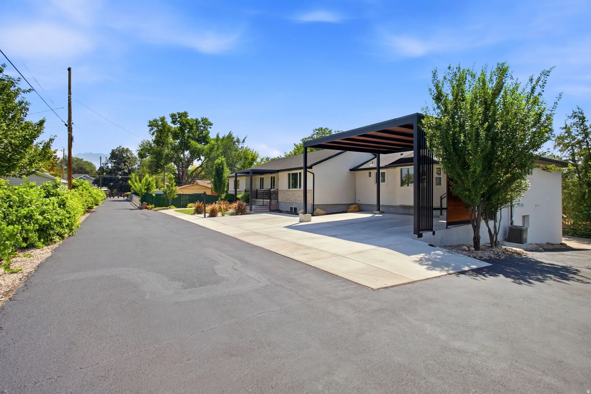 Paved driveway leading to a modern carport with slatted roof, stucco exterior, and mature landscaping