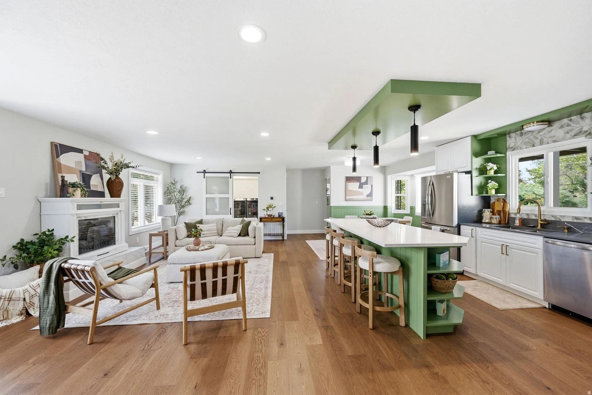 Open-concept living space featuring wood-finish flooring, a white fireplace surround, a kitchen island with seating, pendant lighting, and stainless steel appliances