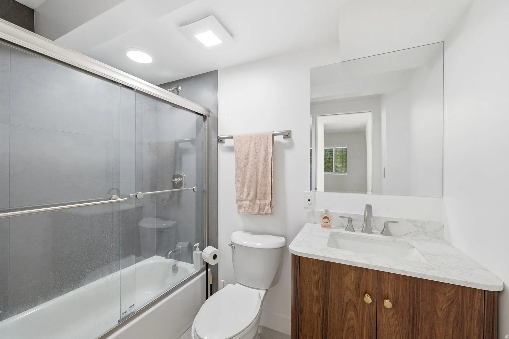 Bathtub with glass sliding doors, dark gray subway tile shower surround, wood-finish vanity with a white countertop, recessed lighting, and a wall-mounted rectangular mirror