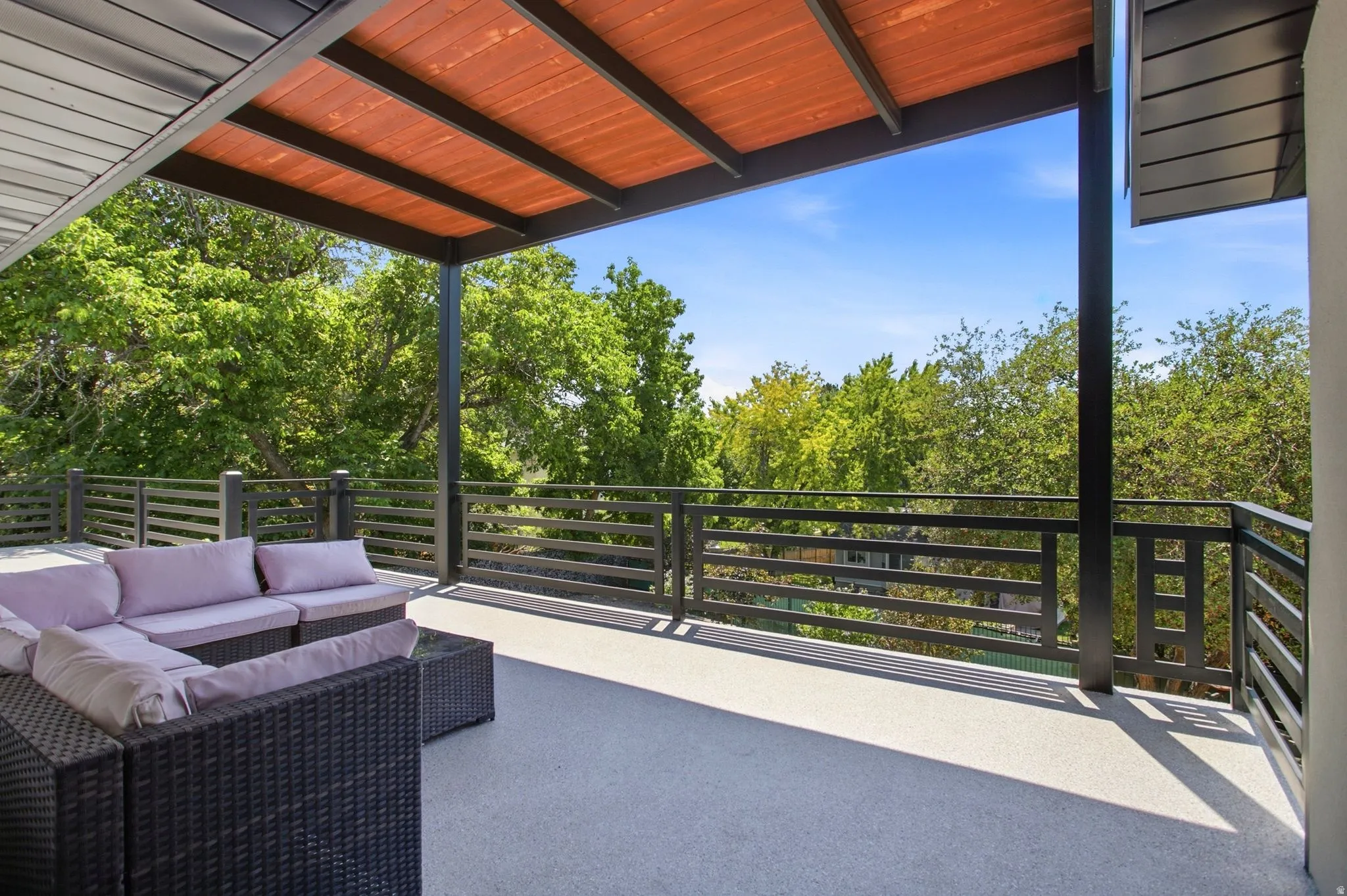 Expansive outdoor deck featuring a textured light gray surface, black metal railings, and a wood-slat overhead covering