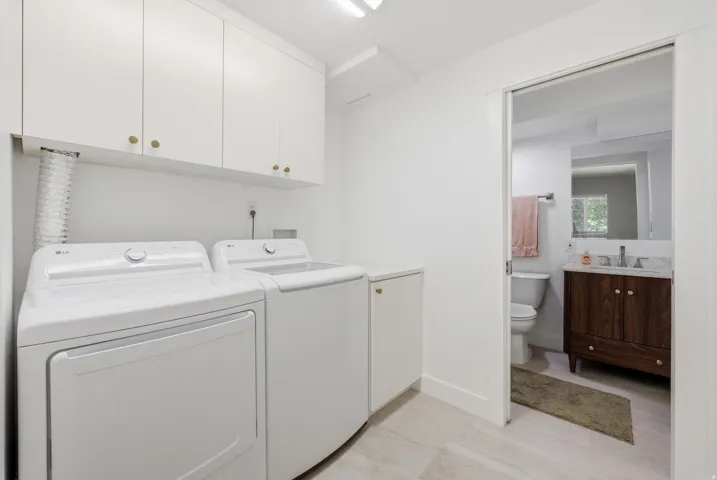 White upper cabinetry with gold-finish hardware, light-toned floor tile, and a sliding pocket door opening to an additional space featuring a wood-finish vanity with a stone countertop